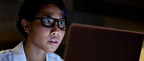 A woman with glasses, using her laptop in the evening to look at 24/7 Virtual Urgent Care telehealth video appointments.