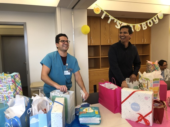 Man standing at table while laughing and opening presents for a baby shower while another man watches and laughs